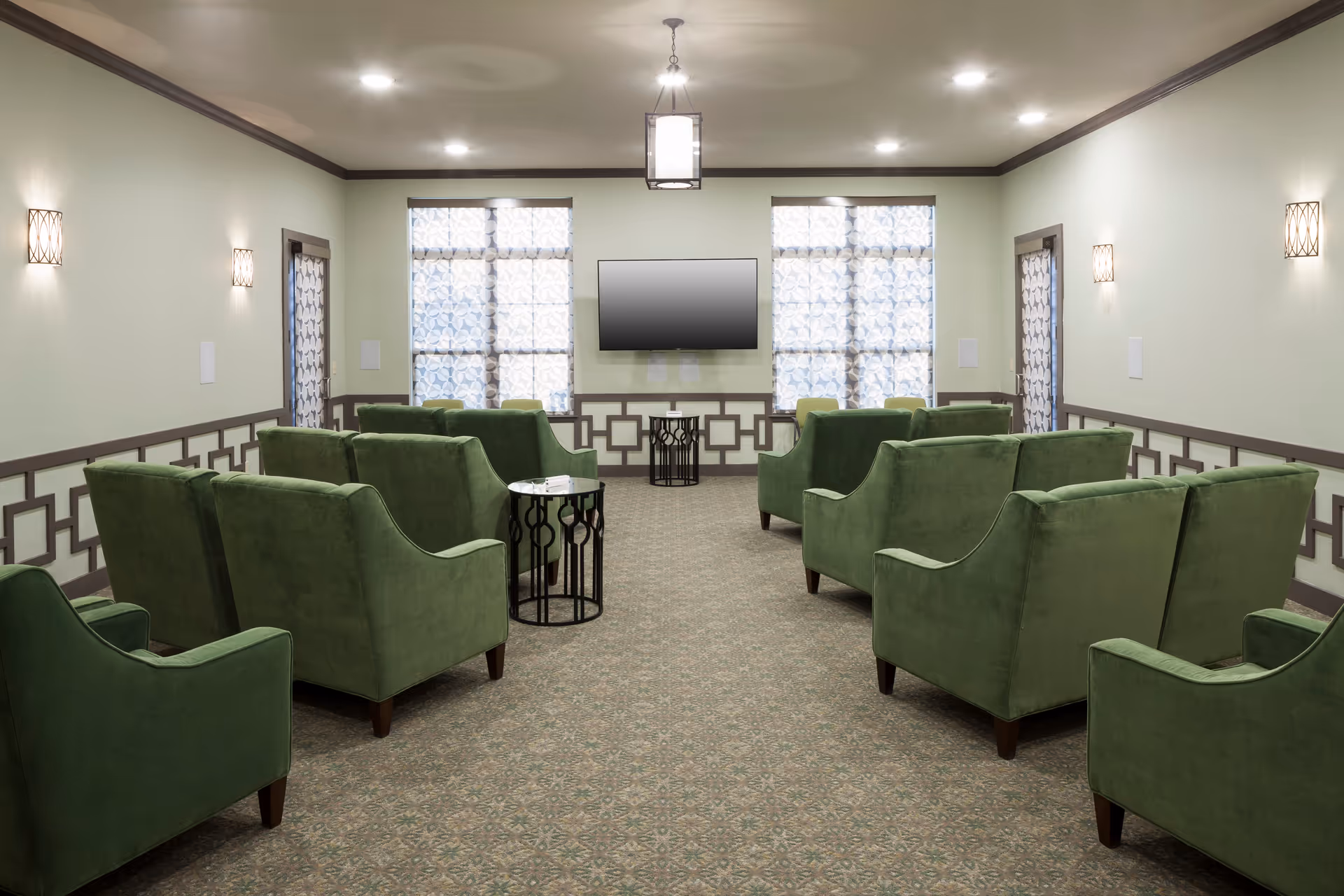 A well-lit common area with green upholstered armchairs arranged in two rows facing a wall-mounted flat screen TV. The room has patterned carpet flooring, light-colored walls with decorative trim, two large windows with patterned curtains, and modern wall sconces and ceiling light fixtures.