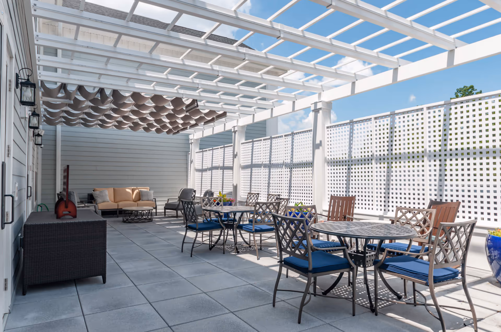 Outdoor patio area with a white pergola overhead, several metal tables with blue cushioned chairs, a beige sofa with gray pillows, and a wicker cabinet. The space is enclosed by white lattice fencing and decorated with large blue planters.