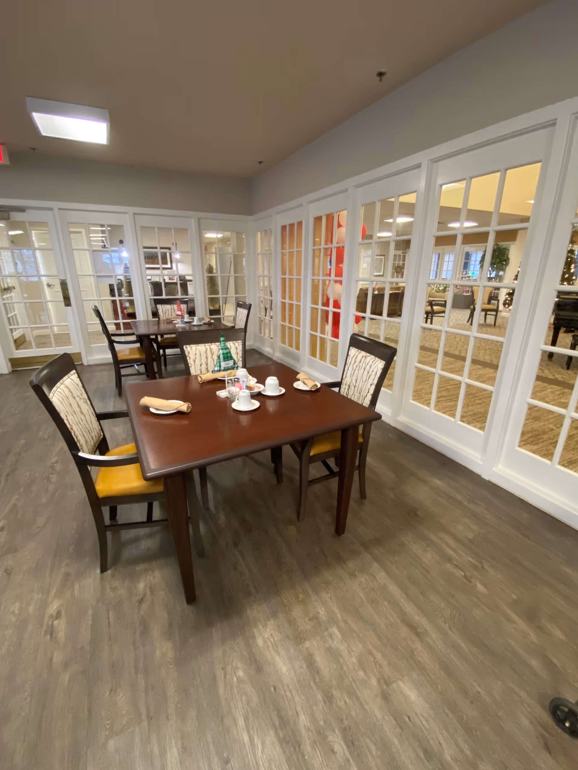 A dining area with wooden tables and chairs set with cups, plates, and napkins. The room has wood flooring and is surrounded by white-framed glass doors and windows, revealing another room with more seating and holiday decorations.