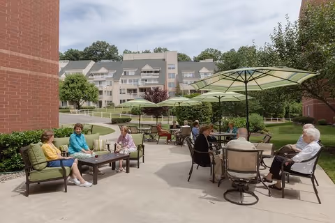 Outdoor patio area at a senior living facility with several elderly people sitting and chatting at tables under large green umbrellas. The background shows a multi-story residential building surrounded by trees and greenery under a partly cloudy sky.