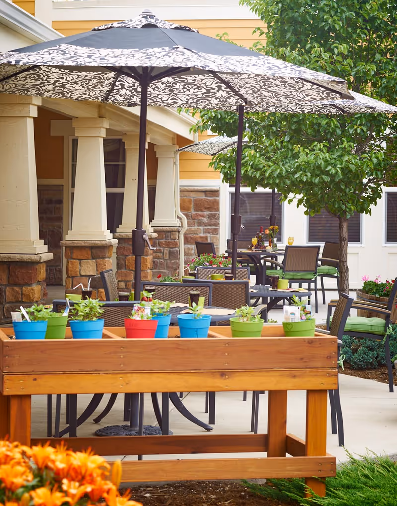 Outdoor patio area at a senior care community with tables and chairs under large patterned umbrellas. A wooden planter box with colorful small pots of plants is in the foreground, and there are trees and flowers around the seating area.