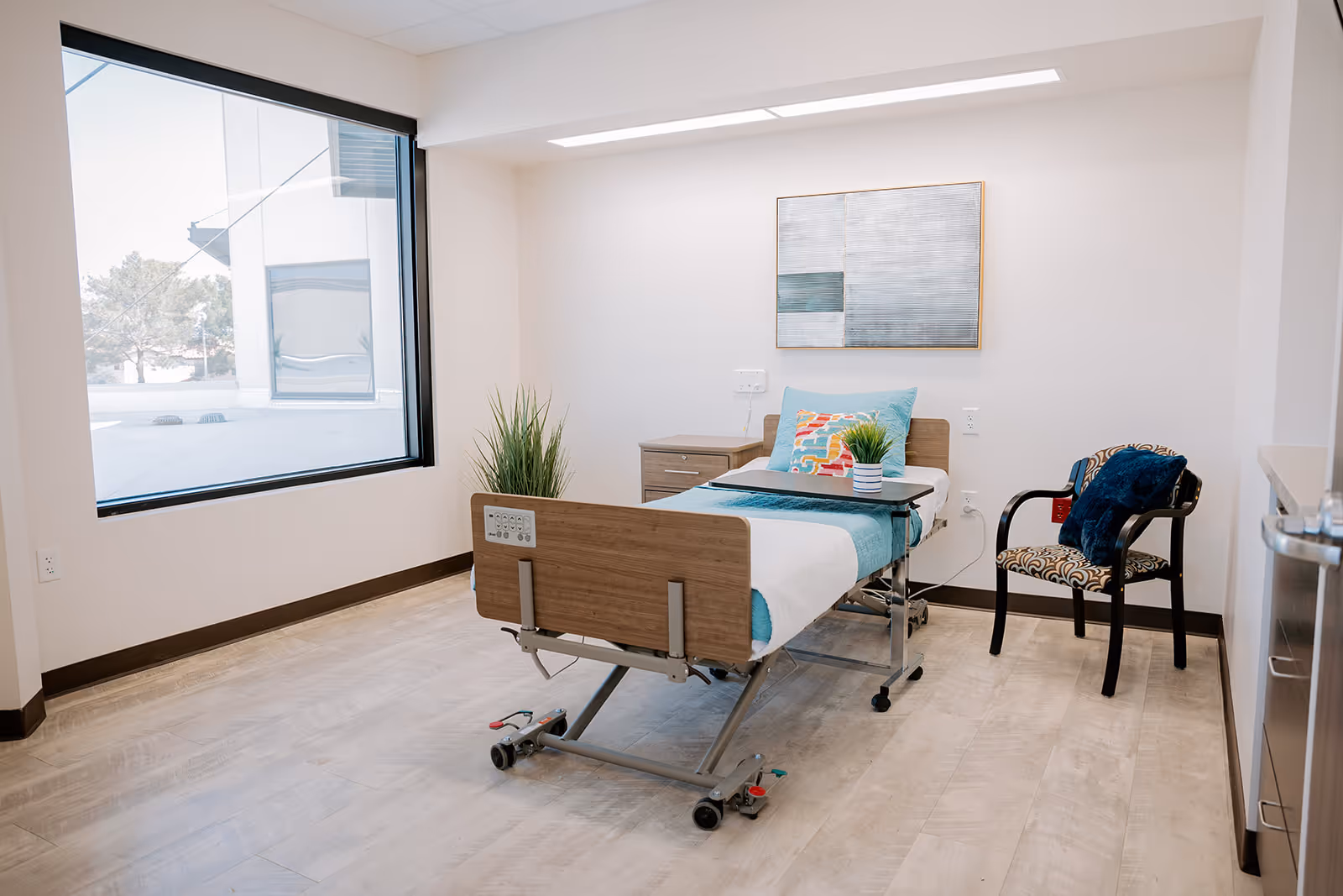 A clean and bright patient room with a hospital bed featuring a colorful pillow and teal blanket. Next to the bed is a small wooden nightstand with a potted plant. A patterned chair with a dark blue cushion is placed near the bed. The room has light-colored walls, a large window letting in natural light, and a modern abstract painting on the wall.