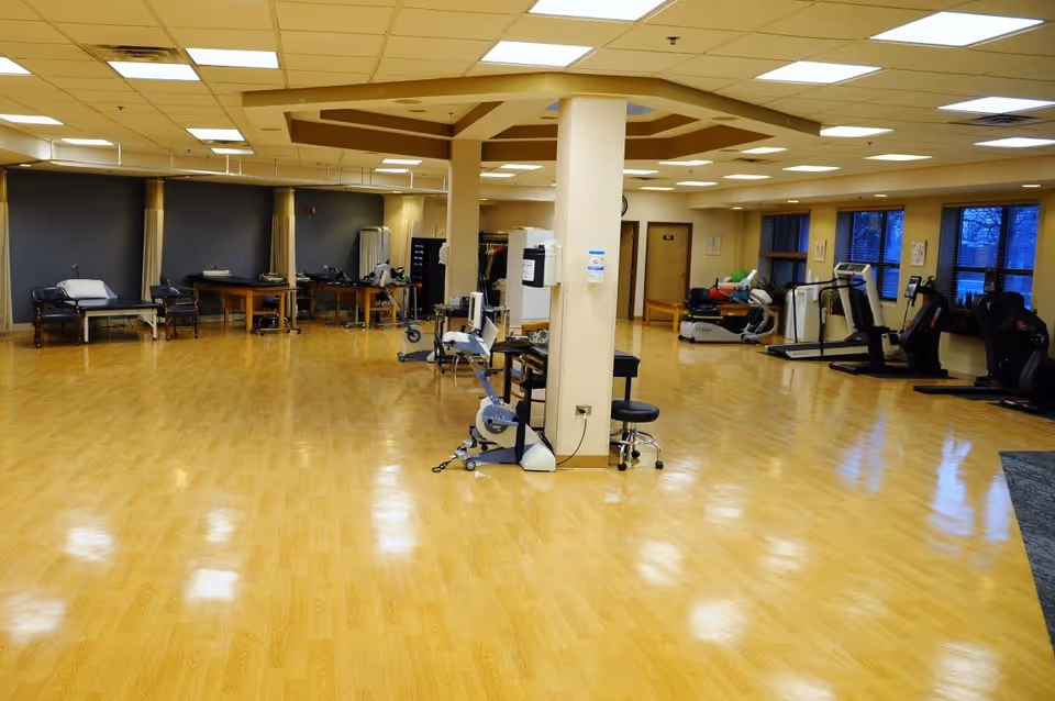A spacious rehabilitation or physical therapy room with wooden flooring, exercise equipment including treadmills and stationary bikes, therapy tables, chairs, and a central pillar. The room has large windows on one side and bright overhead lighting.