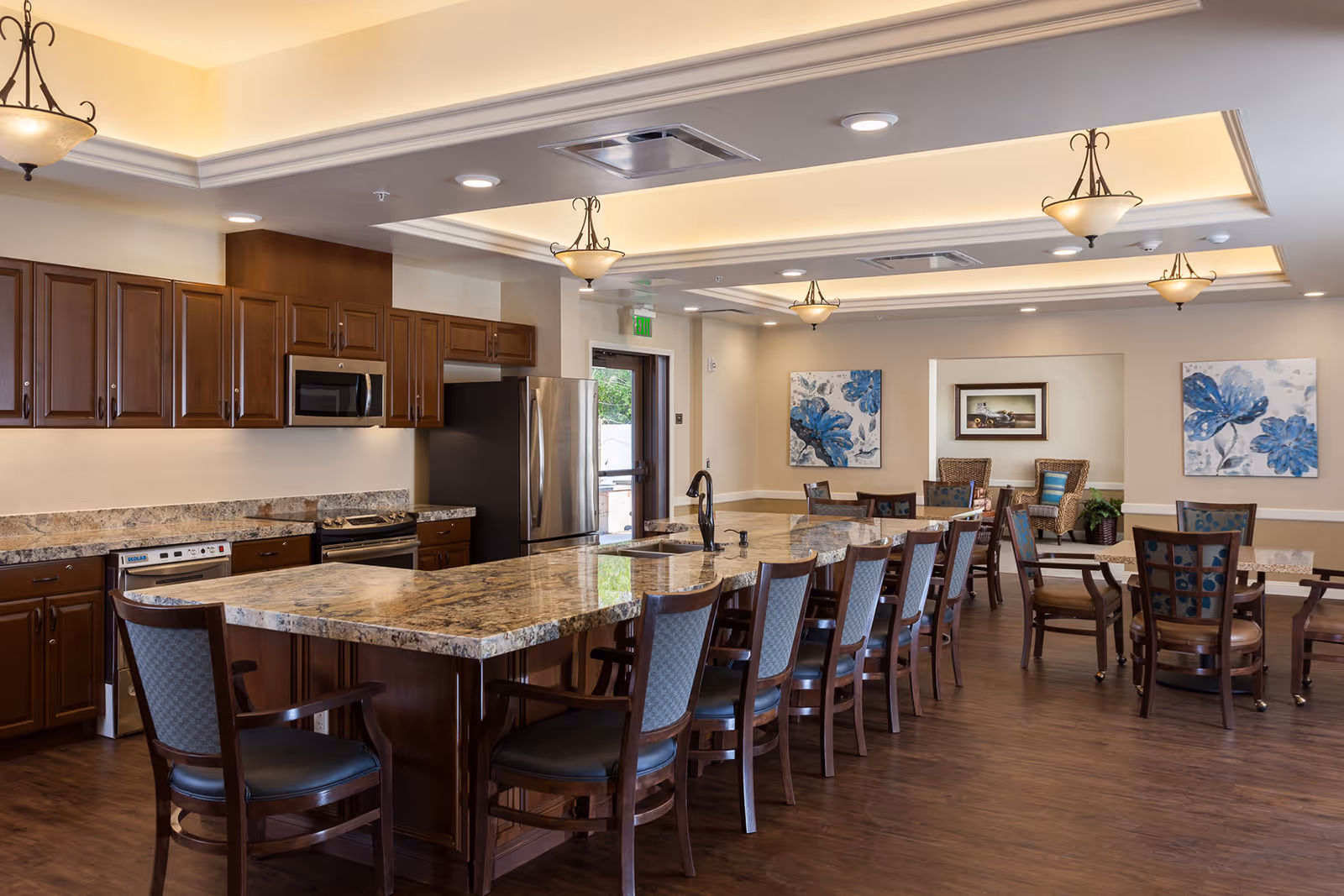 A spacious senior living facility kitchen and dining area with a large marble countertop island surrounded by wooden chairs. The kitchen features dark wood cabinets, a stainless steel refrigerator, microwave, and stove. The dining area has multiple tables and chairs, with blue floral artwork on the walls and warm ceiling lighting fixtures.