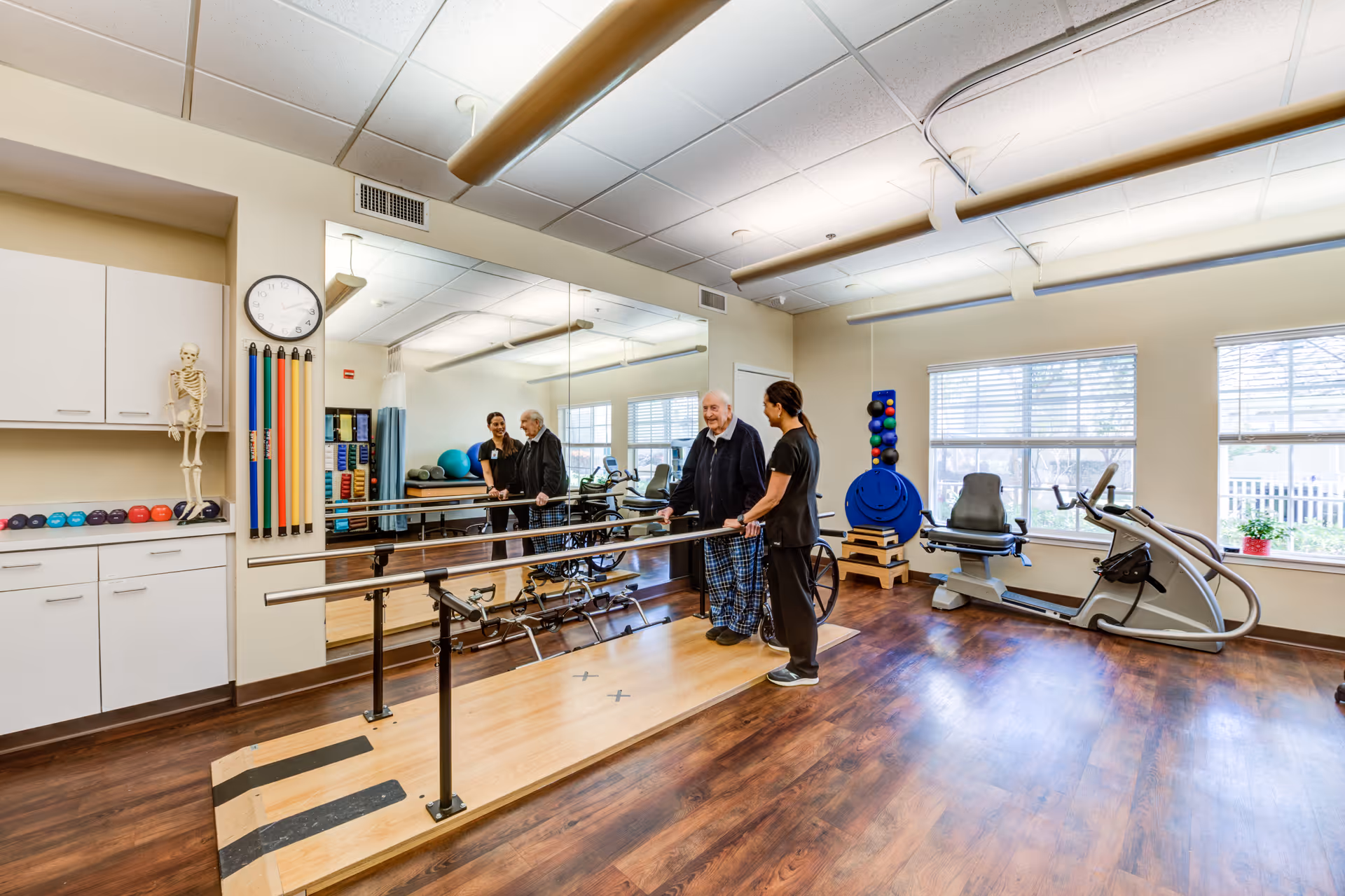 A physical therapy room in a senior living facility with wooden flooring, parallel bars for walking exercises, exercise equipment, and a large mirror on the wall. Two elderly men are standing by the parallel bars, assisted by two caregivers. The room is well-lit with large windows and overhead lights.