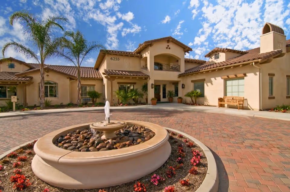 Front entrance of a Mediterranean-style senior living building with a circular fountain, palm trees, and a paved driveway.