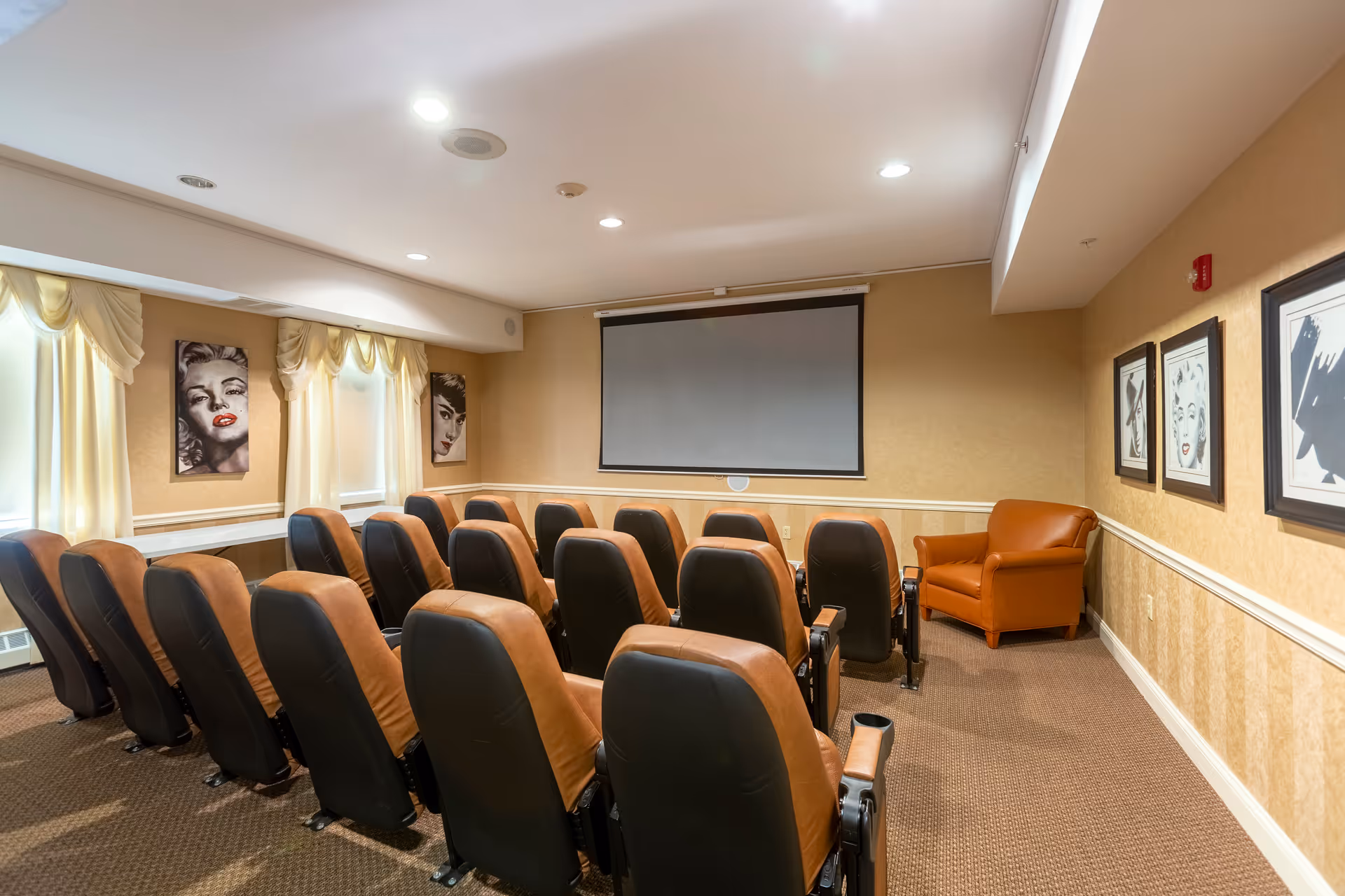 A small theater room with two rows of brown and black cushioned seats facing a blank projection screen. The room has beige walls adorned with framed black and white portraits of classic Hollywood actresses, cream-colored curtains on the windows, and a single brown armchair in the corner.