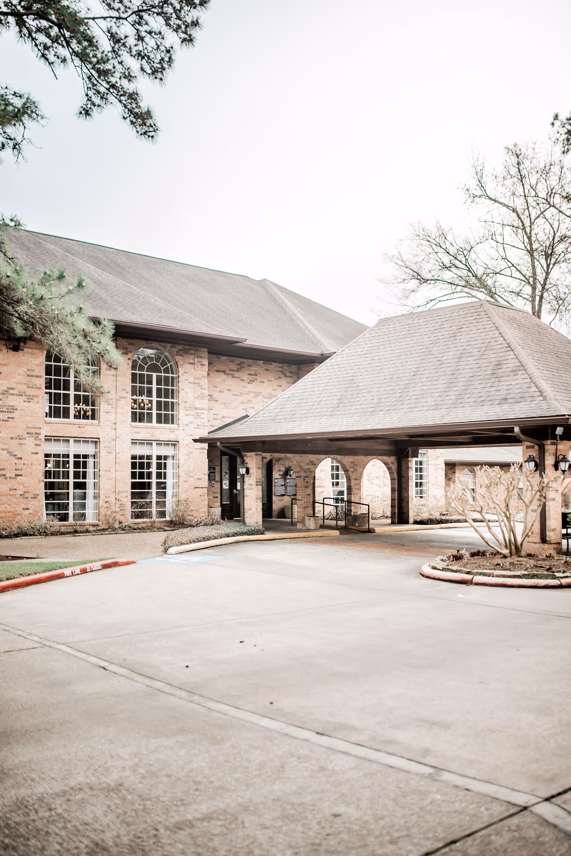 Exterior view of The Woodlands Nursing and Rehabilitation Center showing a brick building with large arched windows and a covered driveway entrance with a peaked roof. Trees and landscaped areas are visible around the building.