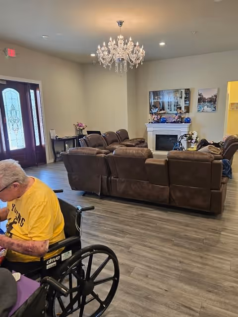 A cozy living room area in a senior living facility with brown leather recliner sofas arranged around a white electric fireplace. A crystal chandelier hangs from the ceiling. There are framed pictures on the wall above the fireplace and a small table with flowers near the entrance door. An elderly person in a yellow shirt is seated in a wheelchair in the foreground.
