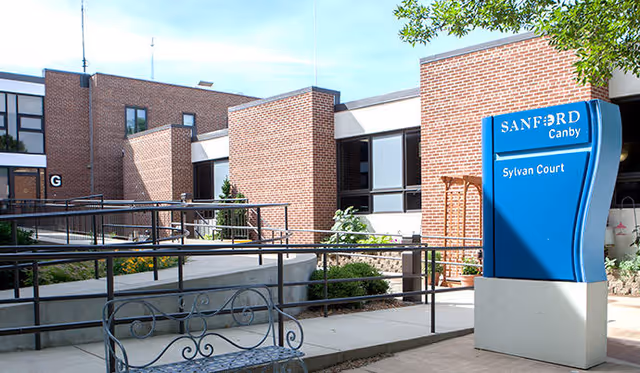 Outdoor courtyard area of Sanford Health Sylvan Court facility featuring brick buildings, a blue sign with the facility name, metal railings, a decorative metal bench, and some greenery under a clear sky.