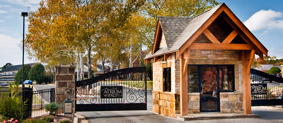 Entrance gate to The Atrium at Serenity Pointe featuring a stone guardhouse with wooden beams and a black wrought iron gate with the facility's name displayed on it. Trees with autumn foliage and a clear sky are visible in the background.