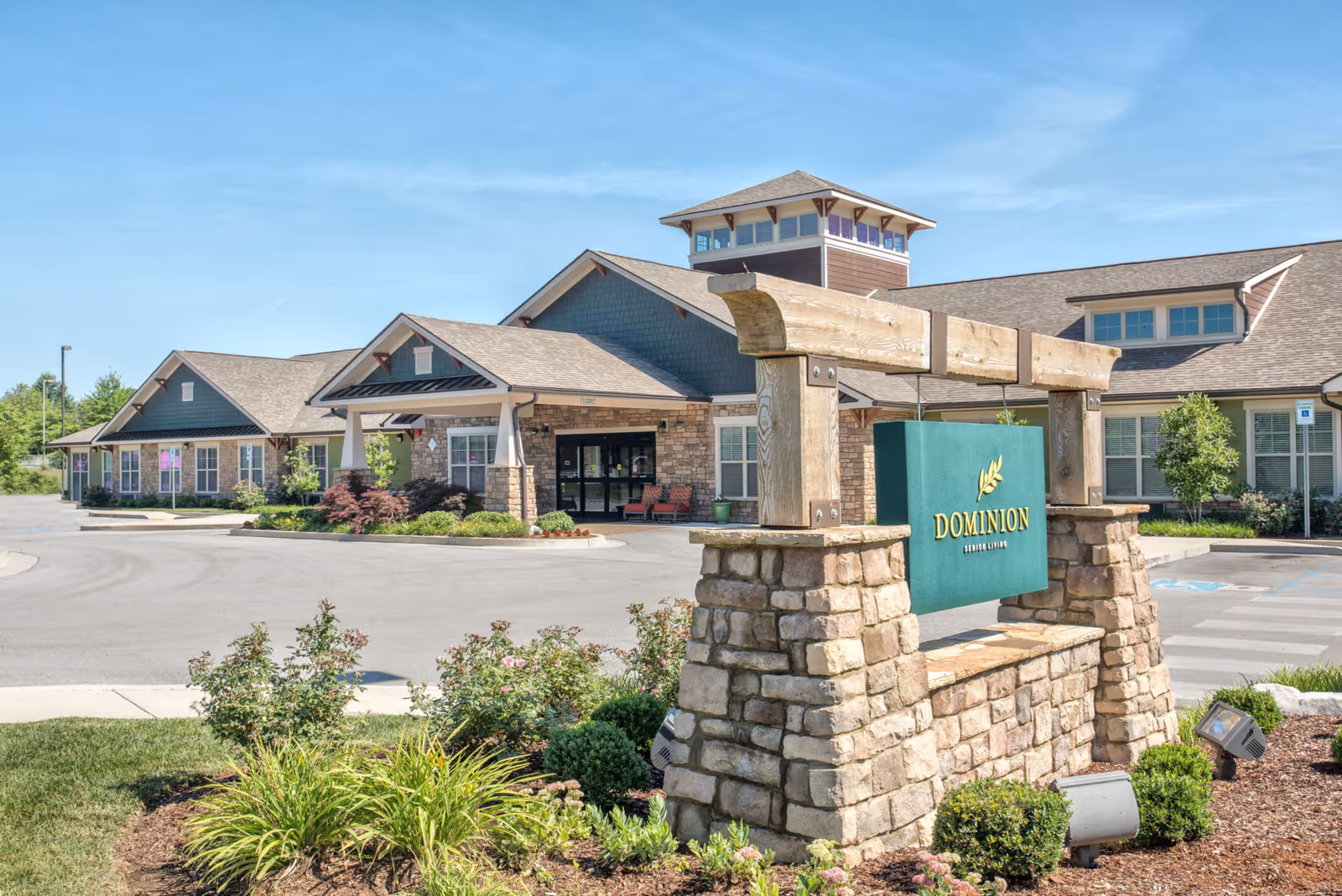 Front exterior of Dominion Senior Living with a stone entrance sign, landscaped driveway, and the facility's building in the background.