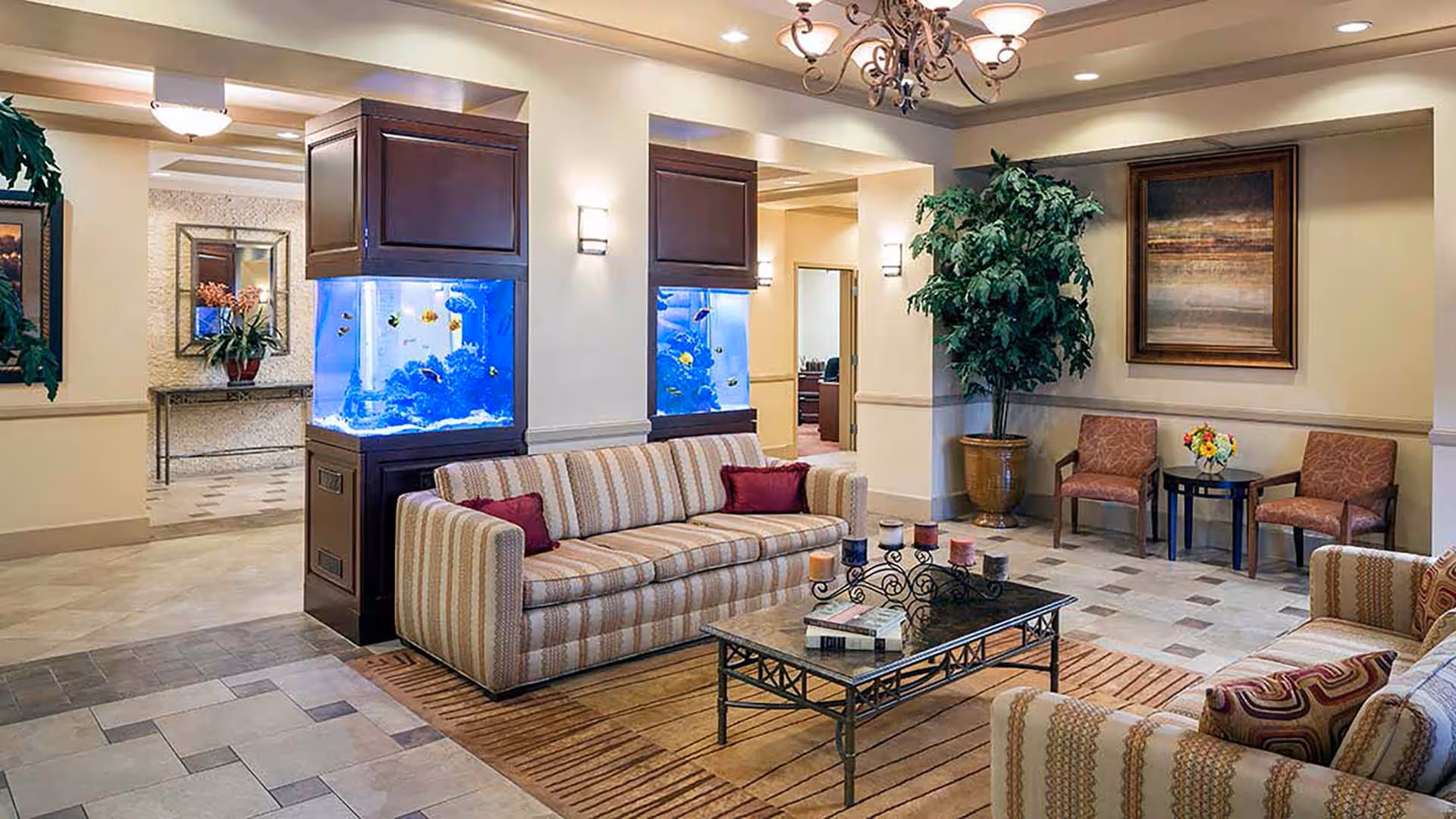 A cozy living room area in a senior living facility featuring two striped sofas with decorative pillows, a glass coffee table with candles and books, two large built-in aquariums with colorful fish, a potted plant, two armchairs with a small table and flower arrangement, wall art, and warm lighting fixtures.