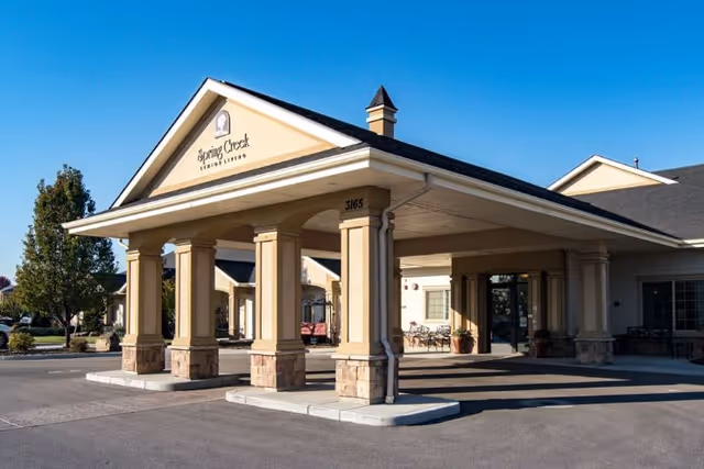 Entrance of a beige senior living building with a covered porte-cochere supported by columns under a clear blue sky.