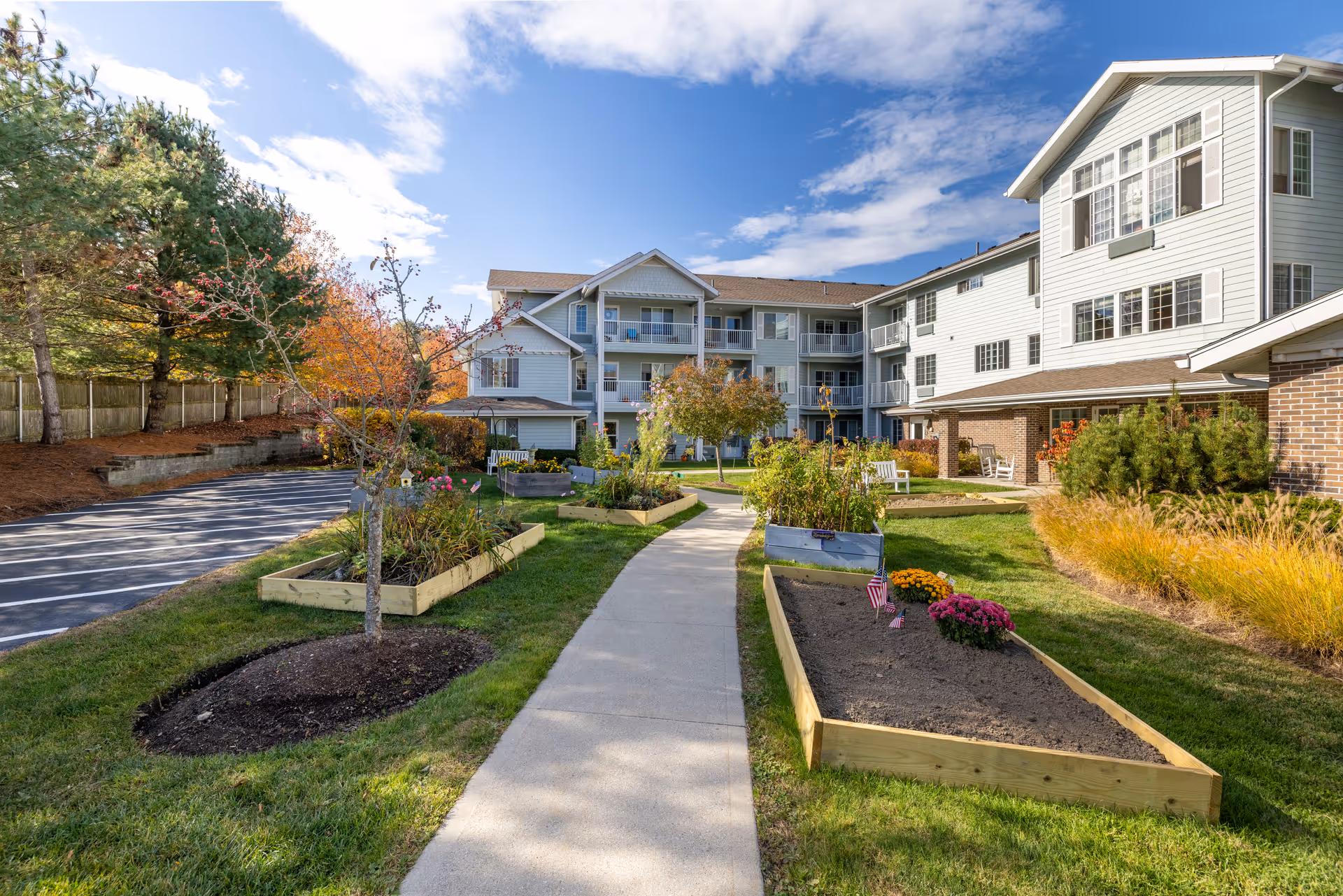 Outdoor garden area at Holiday Kittery Estates with raised garden beds, a paved walkway, trees, and a multi-story residential building in the background under a partly cloudy sky.