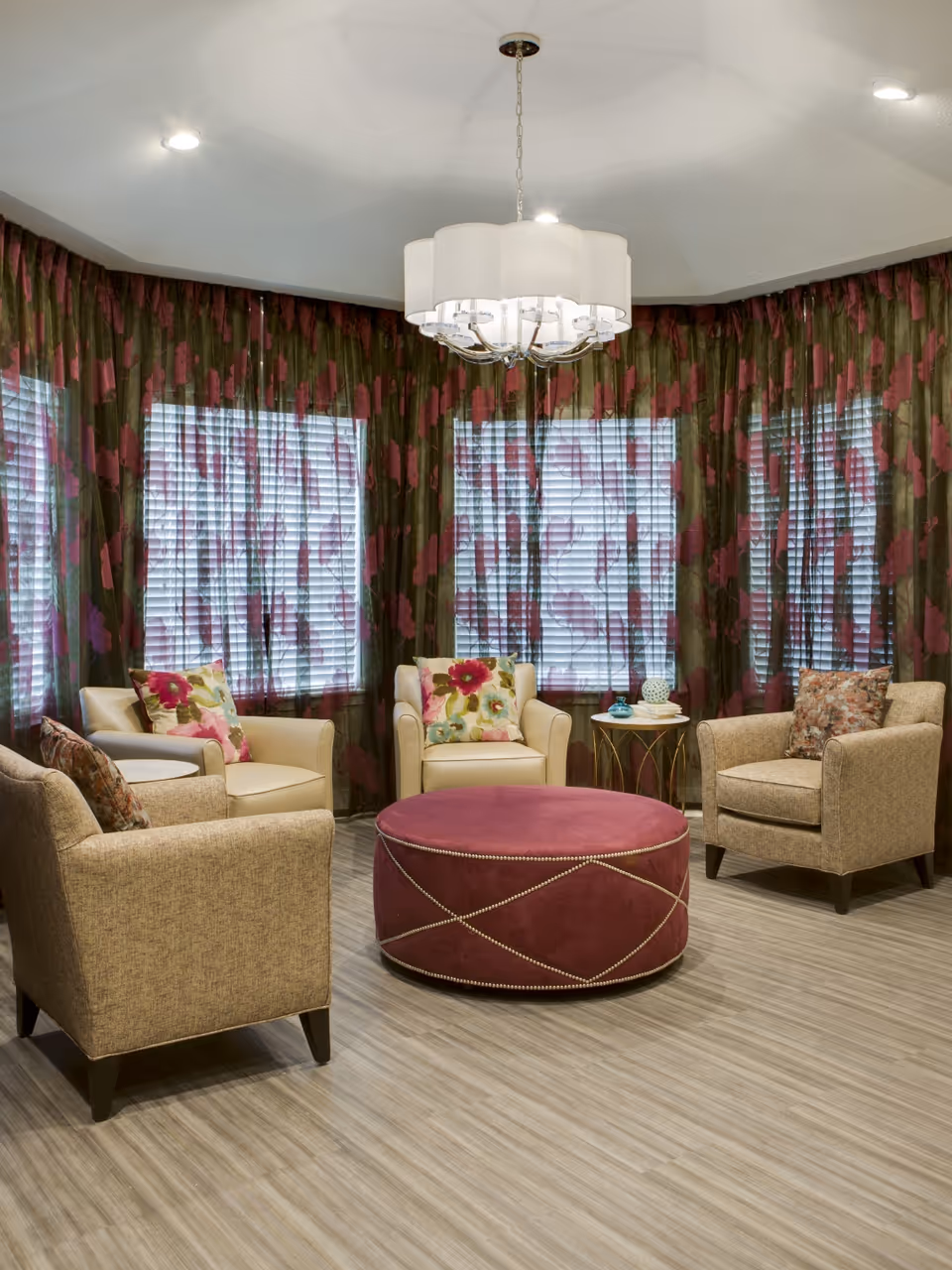 A cozy seating area with four armchairs arranged around a large round pink ottoman beneath a chandelier, in front of floral-patterned curtains and blinds.