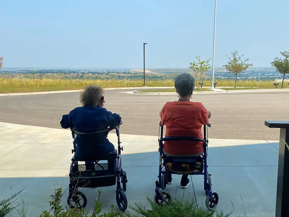 Two elderly individuals sitting on walkers facing away, looking out over a paved area with a scenic view of distant hills and a clear sky.
