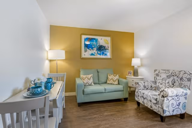 A cozy living room with a light blue loveseat adorned with two patterned pillows, a white and blue patterned armchair, a white side table with a lamp and picture frame, and a small dining table with two chairs set with blue dishes and a flower vase. The back wall is painted mustard yellow and features a colorful abstract painting.