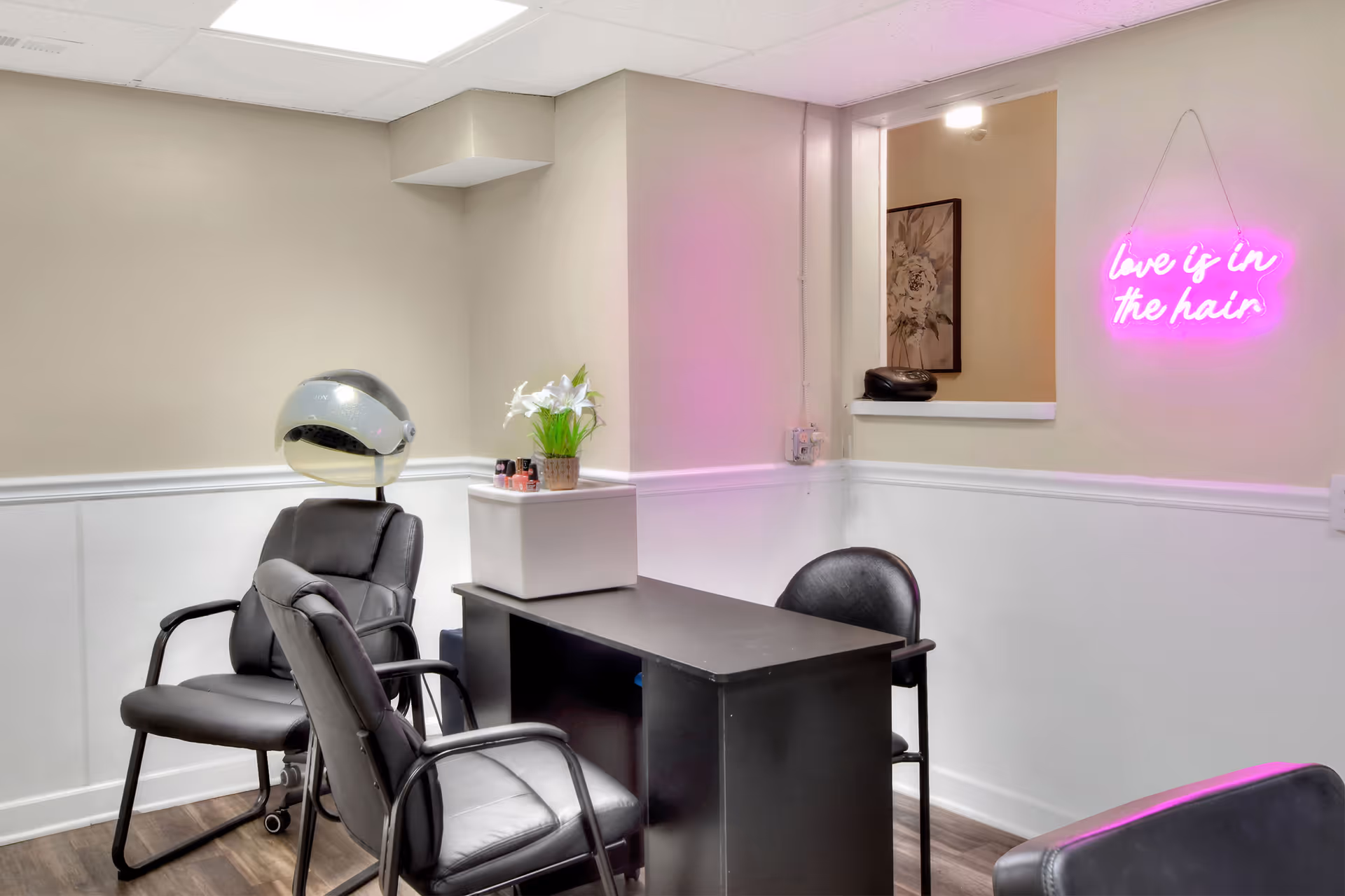 Interior of a small salon or beauty station with black chairs, a hair dryer hood, a black desk, a small white cabinet with flowers and nail polish, and a neon pink sign on the wall that reads 'love is in the hair'.