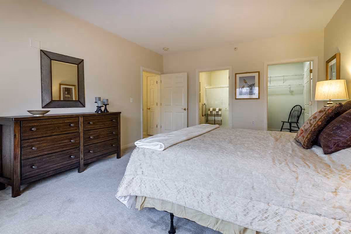 A bedroom with a large bed covered in a light-colored quilt and several pillows. To the left is a dark wooden dresser with a mirror above it and decorative items on top. In the background, there are two open doors, one leading to a bathroom with a glass shower door and the other to a walk-in closet with a black chair inside. A lamp on a bedside table provides warm lighting.