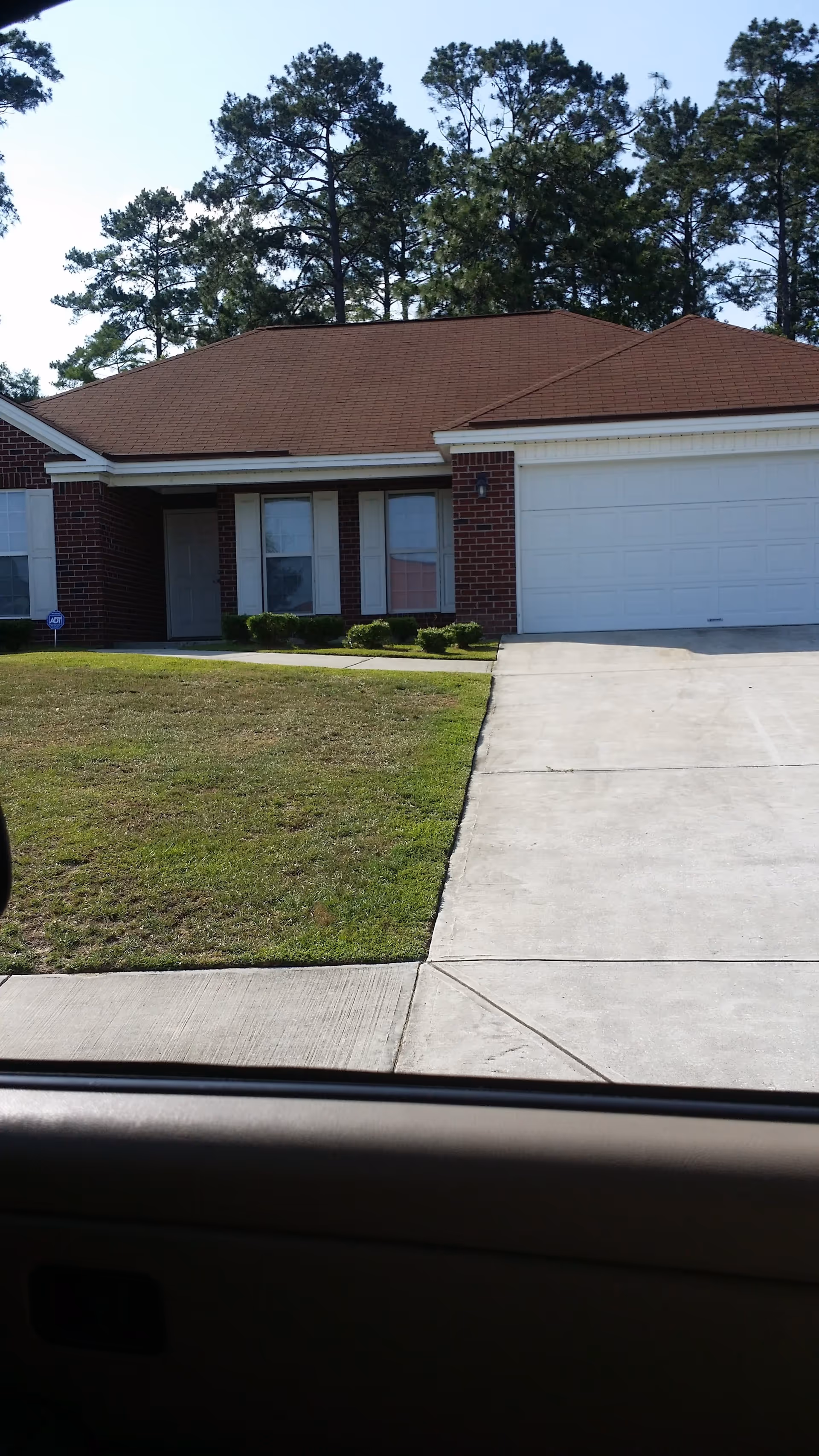 Front exterior view of a single-story brick house with a brown shingled roof, white garage door, and a concrete driveway. There is a small lawn with trimmed bushes in front of the house and tall pine trees in the background. The photo is taken from inside a car, with part of the car door visible at the bottom.