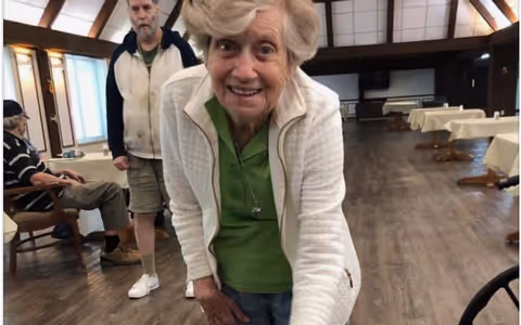 An elderly woman smiles and leans toward the camera in a senior living dining/common room with other residents and tables behind her.