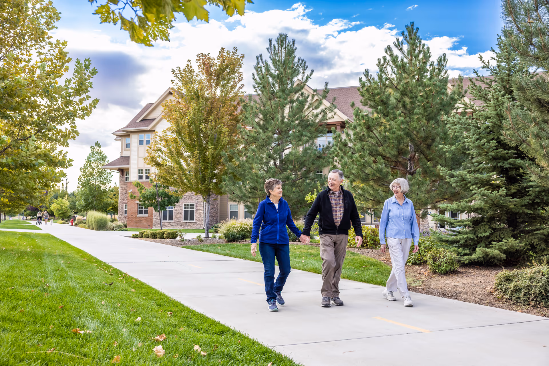 Three elderly people walking and talking on a paved pathway surrounded by green grass, trees, and shrubs with a multi-story residential building in the background under a partly cloudy sky.