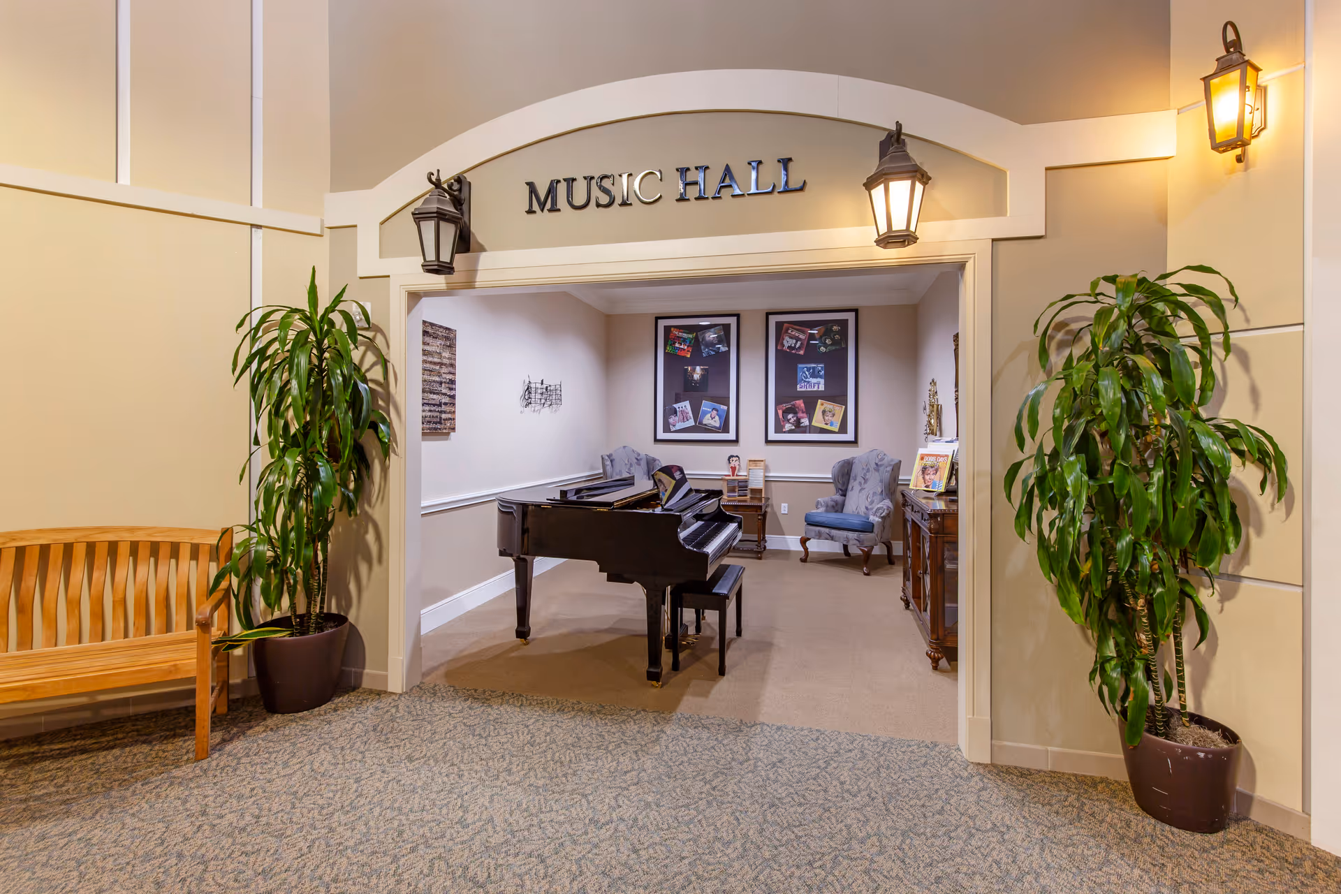 Interior view of a music hall in a senior living facility named Ellery Arbor, featuring a black grand piano with a bench, two upholstered armchairs, framed pictures on the back wall, a wooden side table, and two large potted plants flanking the entrance. The entrance is marked by a sign reading 'MUSIC HALL' with two lantern-style wall lights on either side.