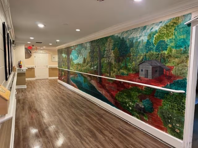 A long indoor hallway with wood-style flooring and beige walls. One side of the hallway features a large colorful mural depicting a nature scene with trees, a river, and a small cabin. The hallway has handrails along the walls and recessed ceiling lights. At the end of the hallway, there is a white door and some wall-mounted fixtures.