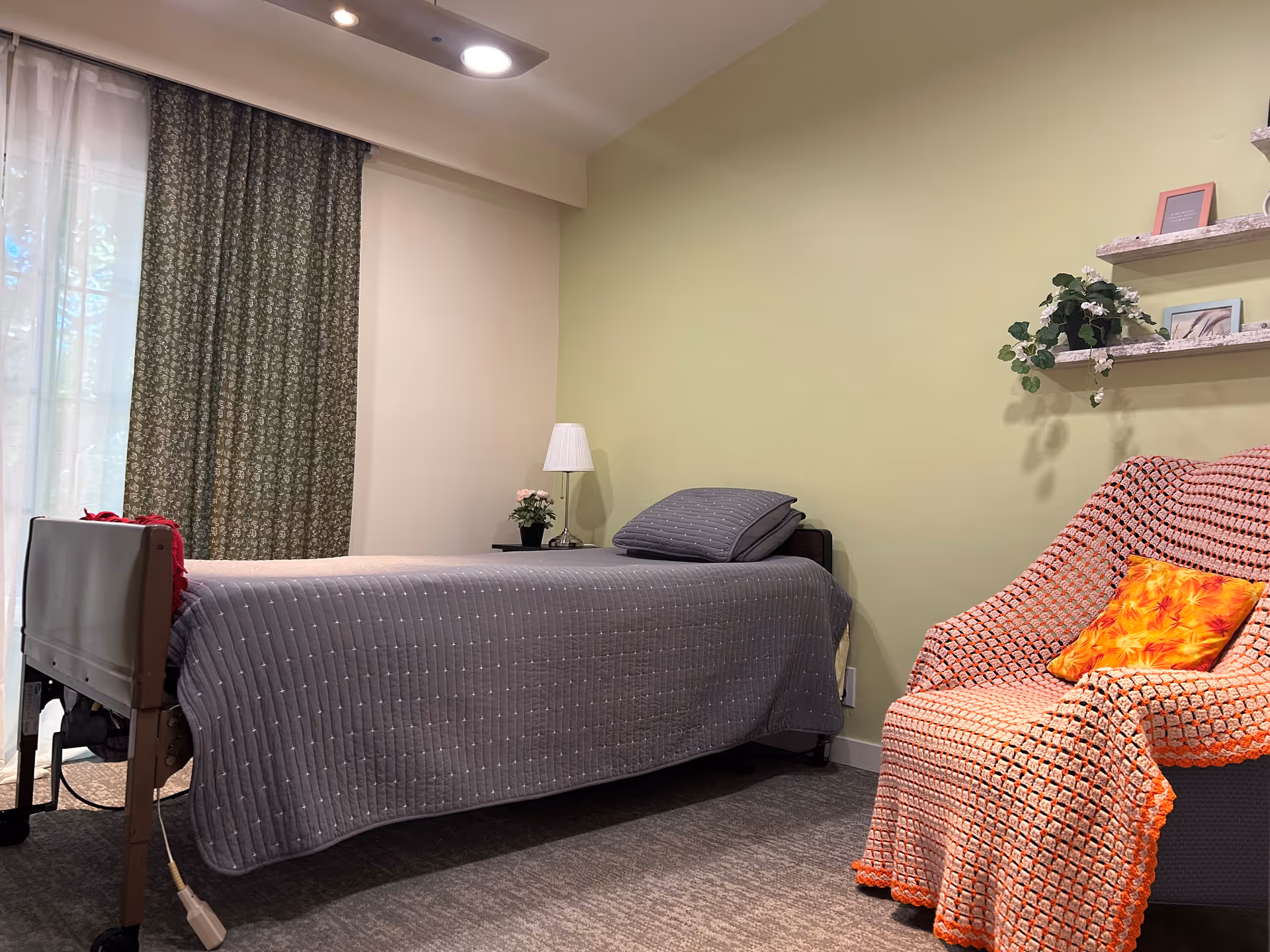 A cozy bedroom in Golden Apricot Manor featuring a single bed with a gray quilt and pillow, a bedside table with a lamp and a small potted plant, a large window with sheer and patterned curtains, and a comfortable armchair covered with an orange crocheted blanket and a bright orange floral pillow. Two small shelves on the wall hold framed pictures and a small plant.
