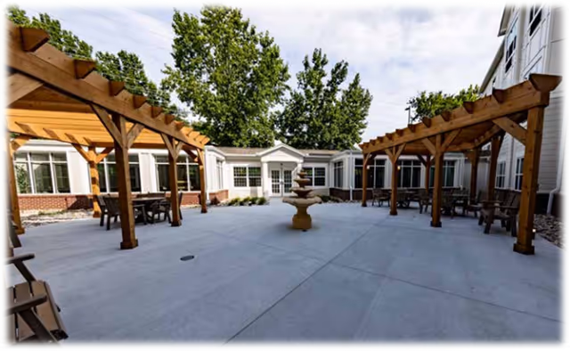 Outdoor courtyard area at Mary Queen of Angels Assisted living facility featuring two wooden pergolas with seating underneath, a central multi-tiered stone fountain, surrounded by building walls with windows and trees in the background.
