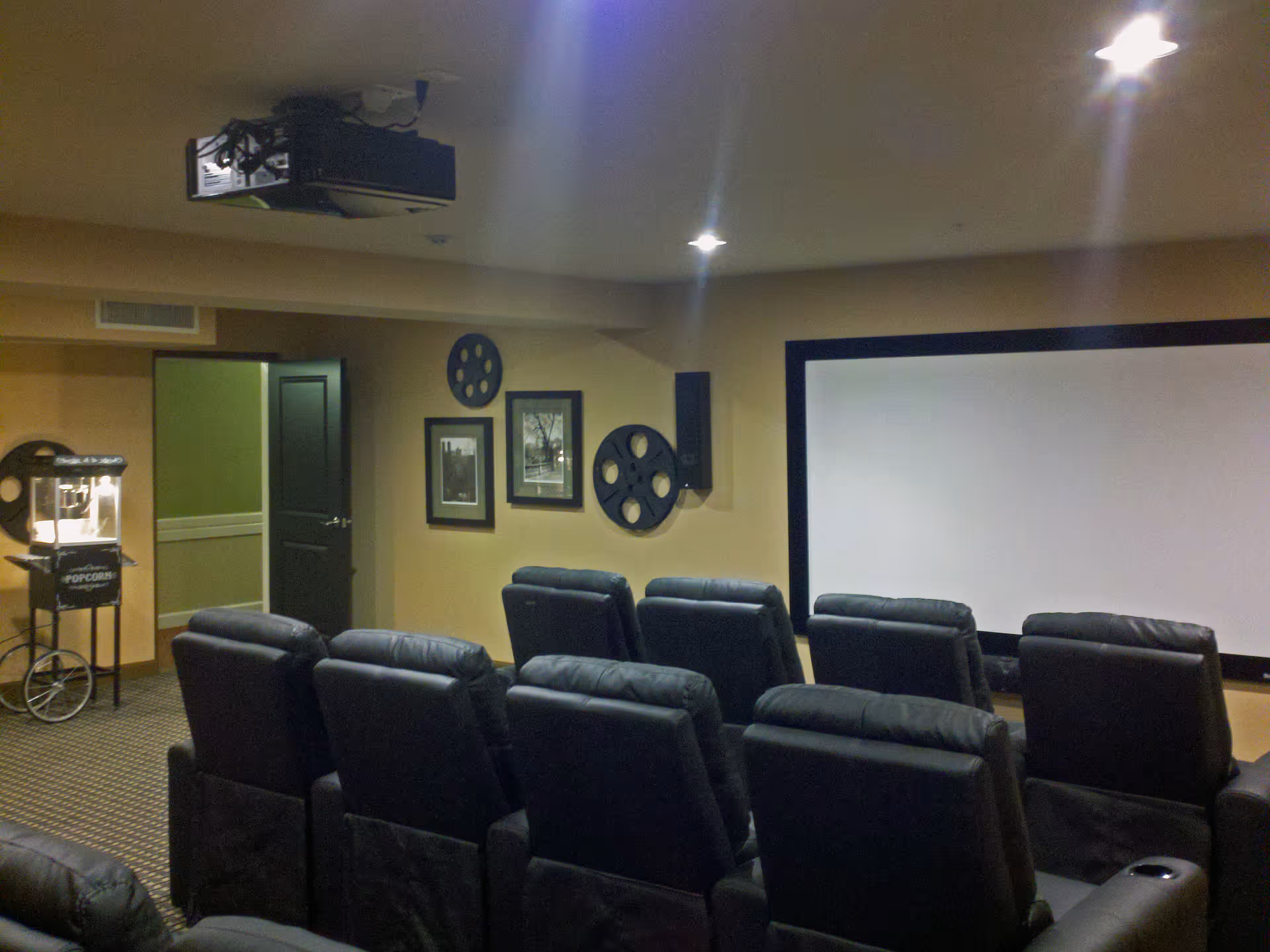 A small movie theater room with two rows of black leather recliner chairs facing a large white projection screen. The walls are decorated with film reel wall art and framed pictures. A popcorn machine is visible near an open door on the left side of the room. Ceiling lights illuminate the space.
