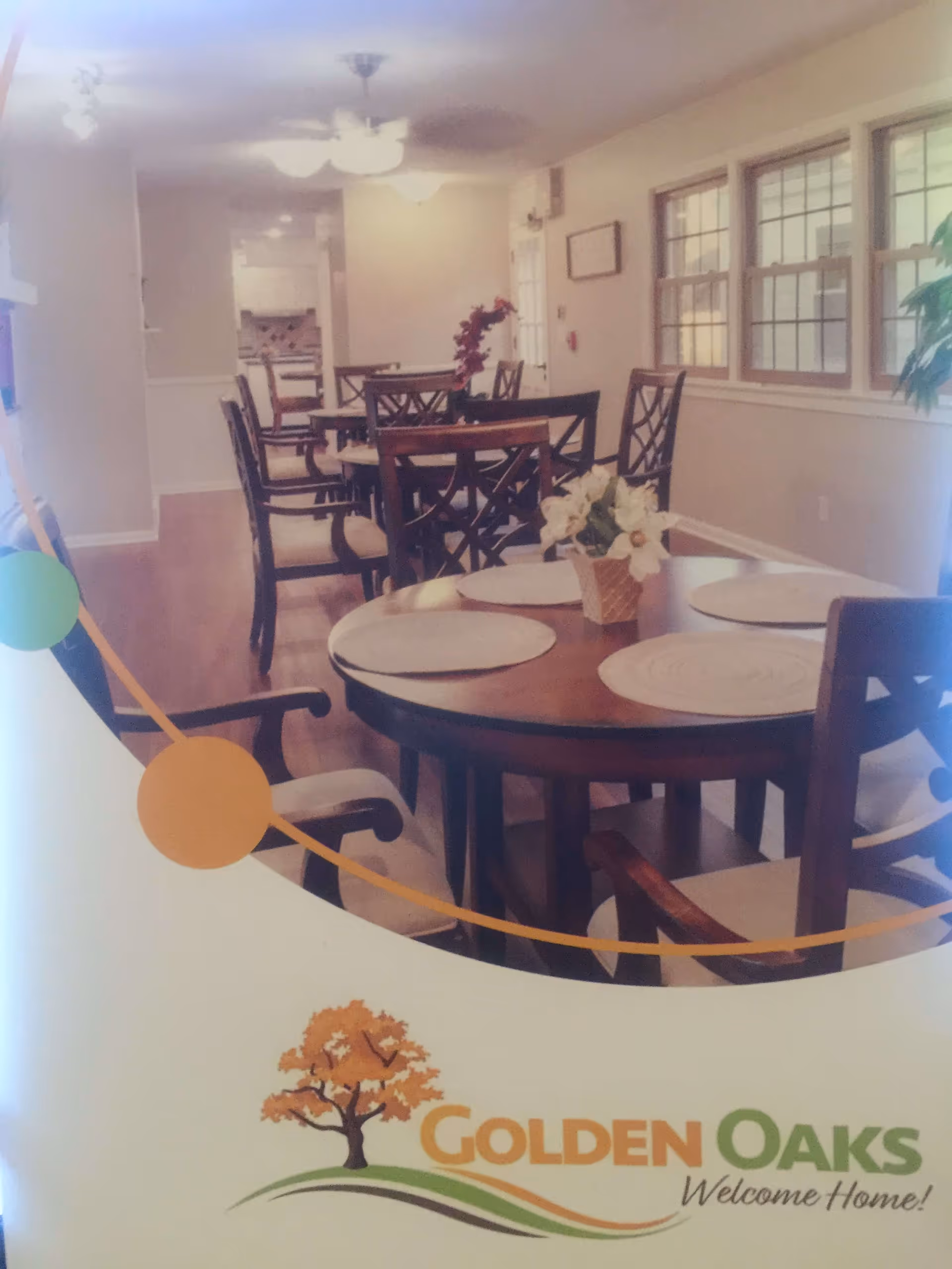 Interior view of a dining room in Golden Oaks Memory Care Home featuring multiple wooden tables with chairs arranged around them. Each table has placemats and a small floral centerpiece. The room has large windows on one side, light-colored walls, and ceiling fans with lights.