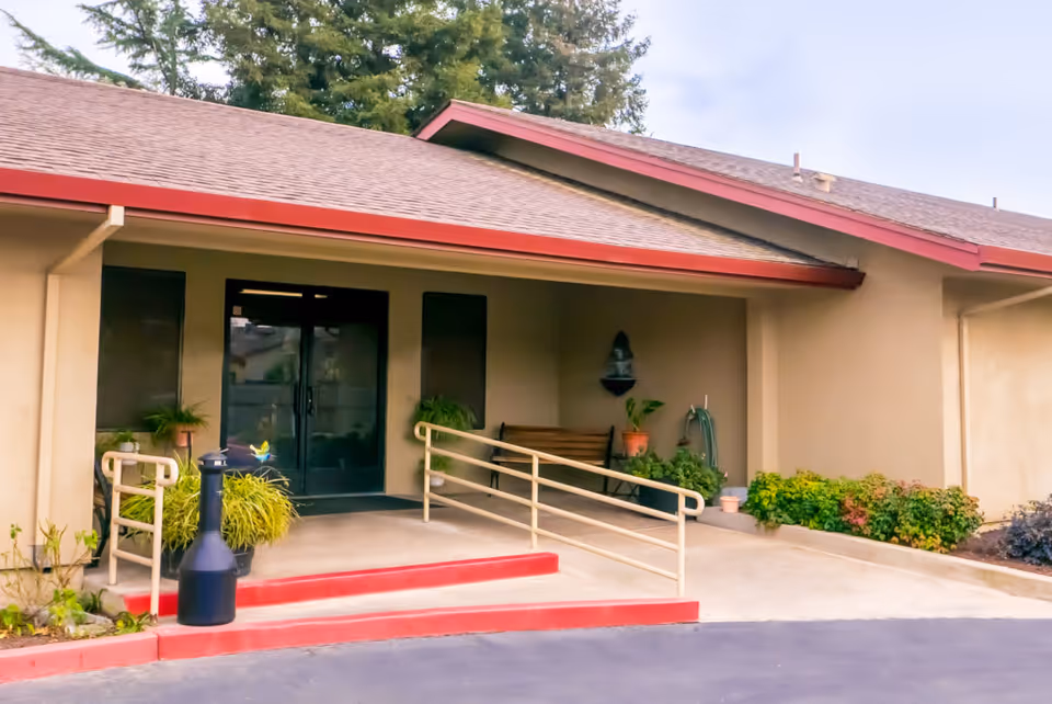 Entrance to a building with a covered porch, double glass doors, a wheelchair ramp with handrails, potted plants, a bench, and a small wall-mounted fountain. The building has a beige exterior with a red trim roof and surrounding greenery.