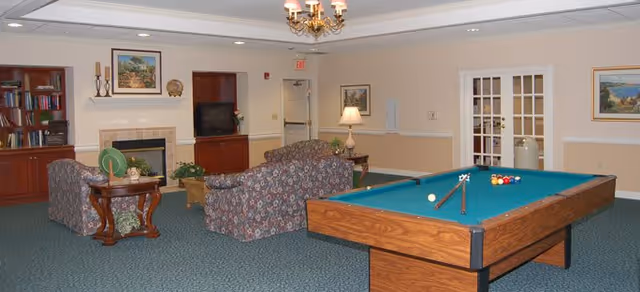 A cozy common room featuring a pool table with billiard balls and cues, two floral-patterned sofas facing a fireplace with a TV on a wooden cabinet, a side table with a lamp, bookshelves, and framed artwork on the walls.