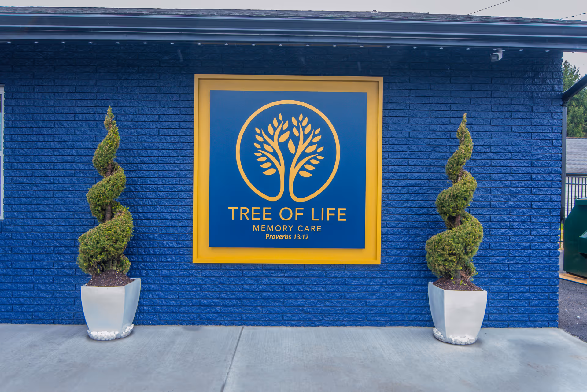 Exterior wall of a building painted blue with a large square sign featuring a yellow tree logo and the text 'TREE OF LIFE MEMORY CARE Proverbs 13:12'. Two spiral-shaped topiary plants in white pots are placed symmetrically on either side of the sign.