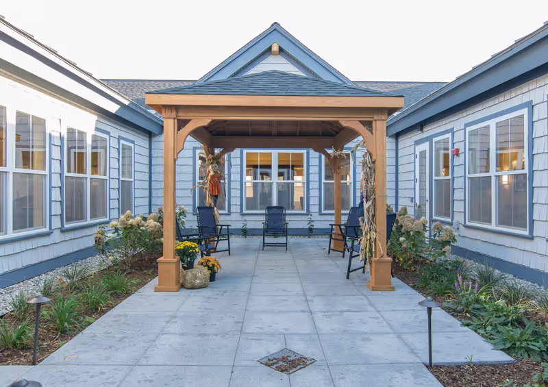 Outdoor courtyard area at a senior living facility featuring a wooden pergola with four black chairs underneath. The courtyard is surrounded by blue-gray buildings with multiple windows and landscaped garden beds with plants and flowers on either side of the paved walkway.
