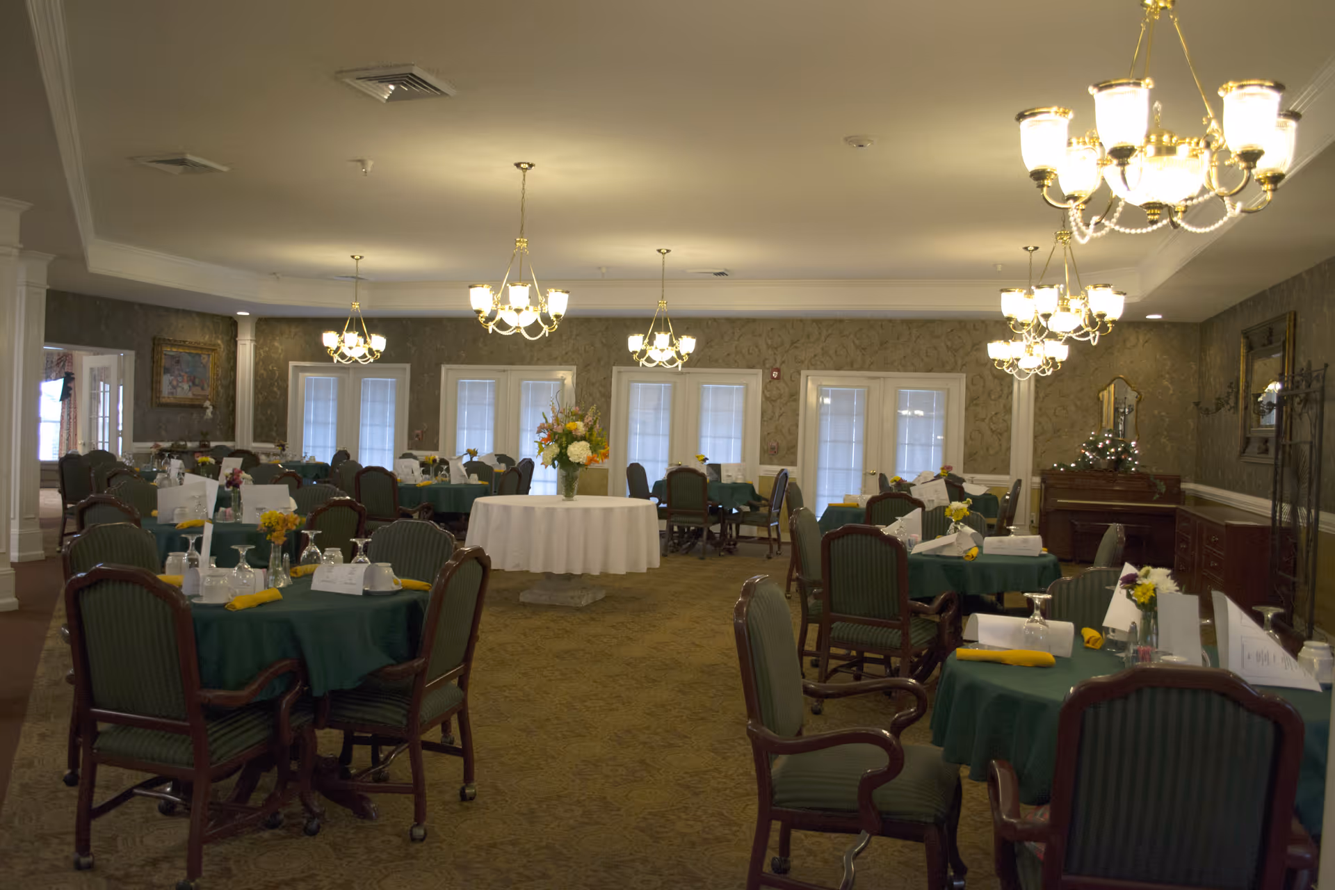 A spacious dining room with multiple round tables covered in green tablecloths, each set with cups, glasses, napkins, and small flower arrangements. The room is well-lit with several chandeliers hanging from the ceiling. There is a large round table with a floral centerpiece in the middle of the room. The walls have a patterned wallpaper, and there are several windows with white blinds.