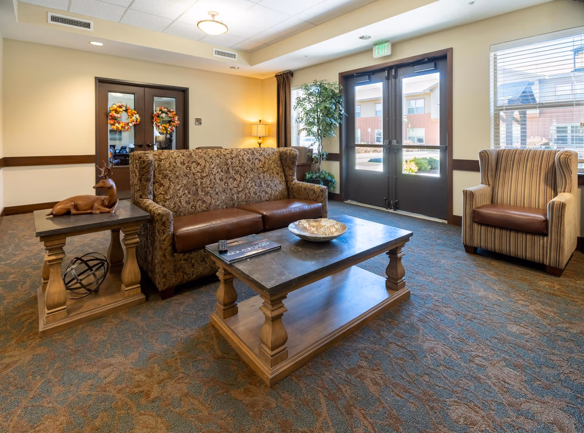 A cozy senior living facility lounge area with a patterned sofa, a striped armchair, a wooden coffee table with a decorative bowl, and a side table with a deer figurine. The room has carpeted flooring, double glass doors with autumn wreaths, a large window, and a potted plant near the exit door.