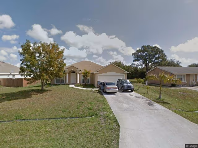 Front view of a single-story suburban house with a driveway and two cars on a grassy lawn under a blue sky.