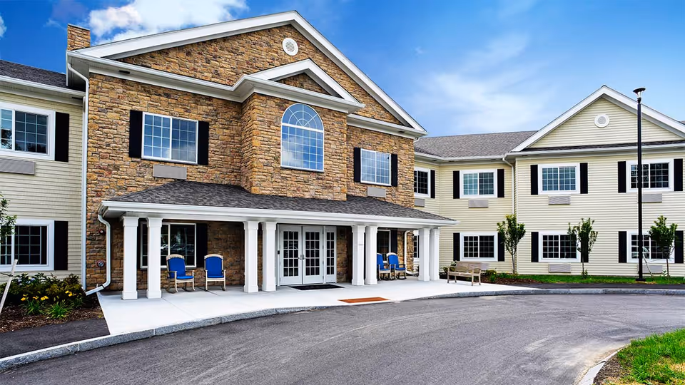 Front entrance of a two-story senior living building with a stone facade, covered porch with chairs, and a curved driveway.