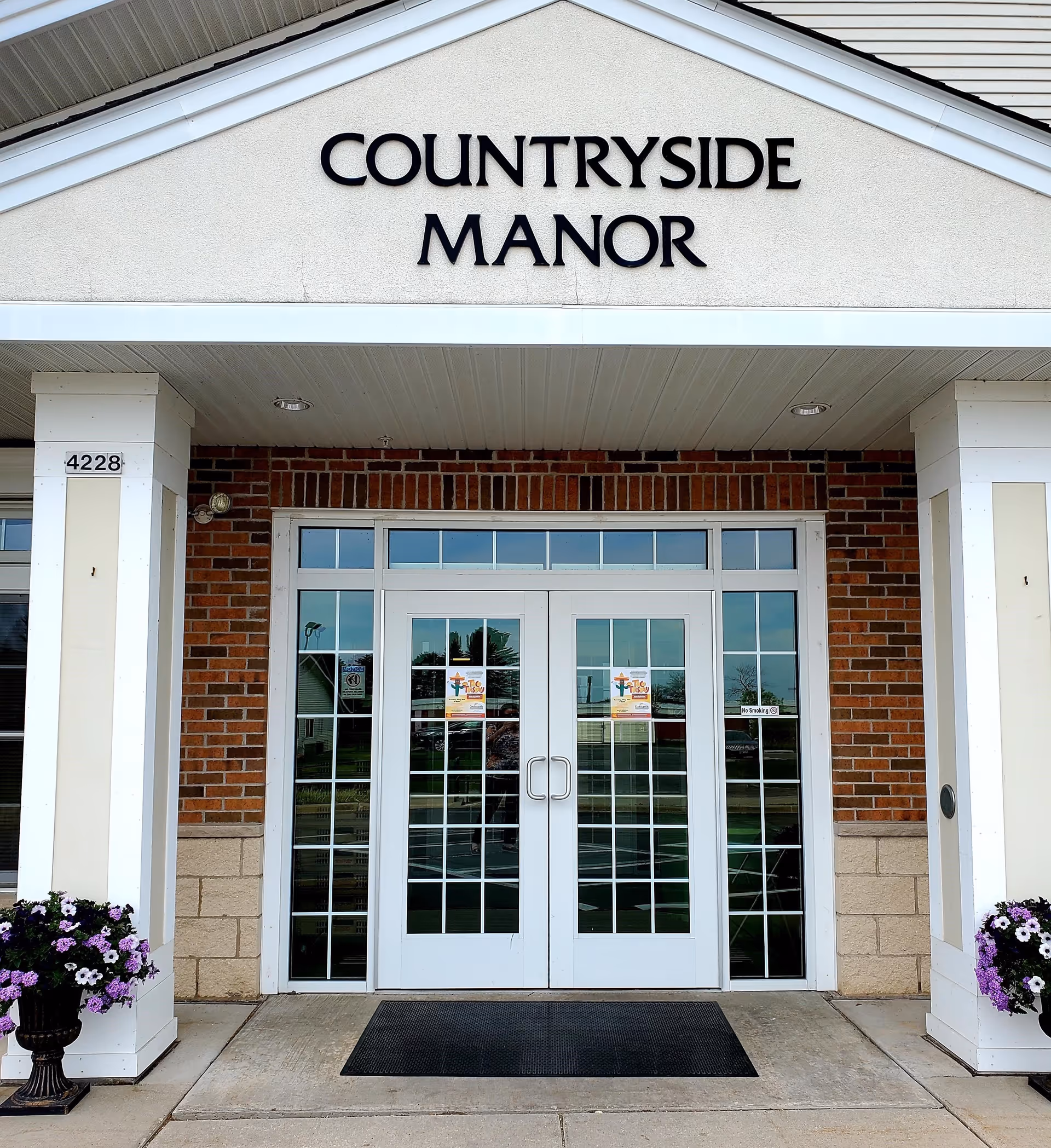 Entrance to Countryside Manor with double glass doors framed by brick and white columns, two flower pots with purple and white flowers on either side, and the address number 4228 displayed on the left column.