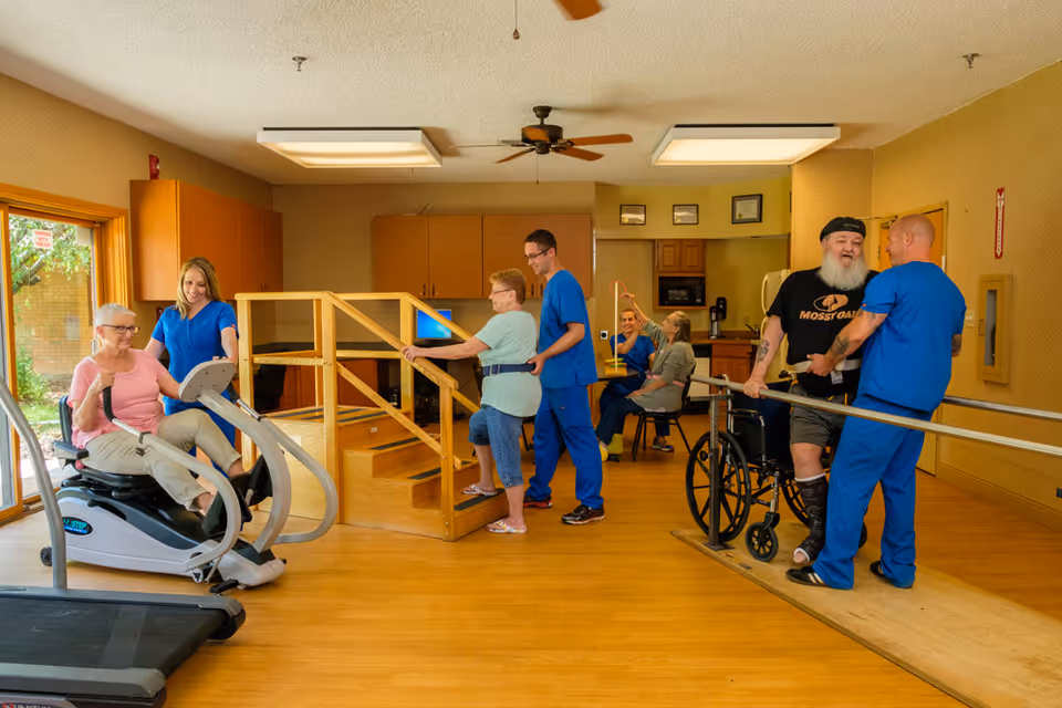 A rehabilitation room with elderly patients and healthcare workers engaging in physical therapy exercises. One elderly woman is using a seated exercise machine with assistance from a female healthcare worker. Another elderly woman is walking up wooden steps with support from a male healthcare worker. A man with a beard and a black hat is practicing walking with parallel bars, assisted by another male healthcare worker. In the background, two women are seated at a table, one of whom is holding a yellow exercise tool. The room has wooden floors, ceiling fans, and cabinets along the walls.