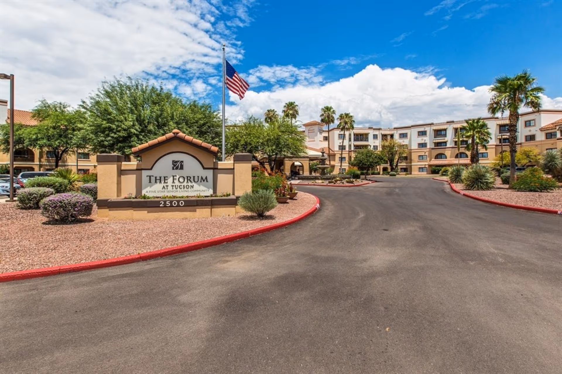 Exterior view of The Forum at Tucson senior living community with a large sign at the entrance, an American flag on a flagpole, landscaped bushes and trees, and a multi-story building in the background under a partly cloudy blue sky.