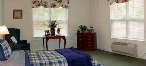 A furnished senior living bedroom with a plaid bedspread, dresser, window, and plants.