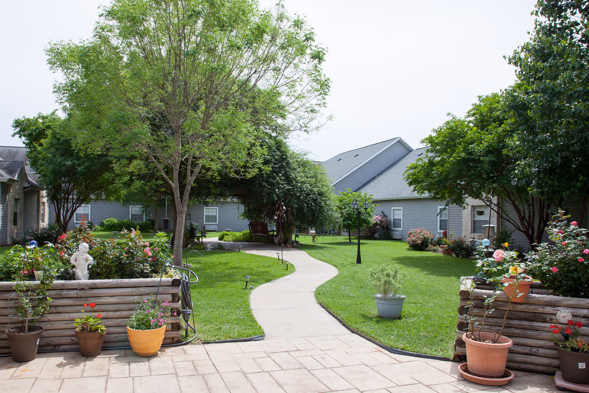 A landscaped courtyard with a winding sidewalk, potted plants, trees, and single-story buildings around.