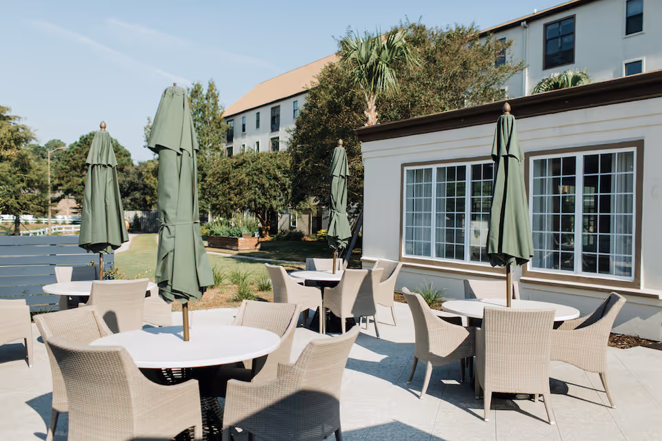 Outdoor patio area with round tables and wicker chairs, each table having a closed green umbrella. The patio is adjacent to a building with large windows and surrounded by trees and greenery under a clear sky.