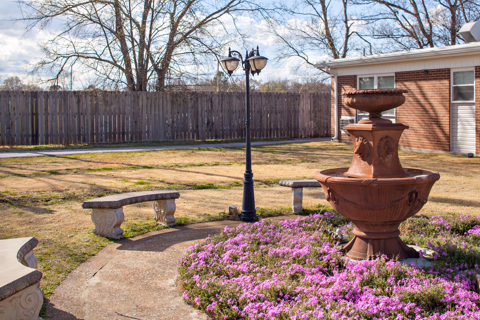 Outdoor courtyard featuring a multi-tiered stone fountain surrounded by purple flowers, stone benches, and a lamppost beside a brick building.