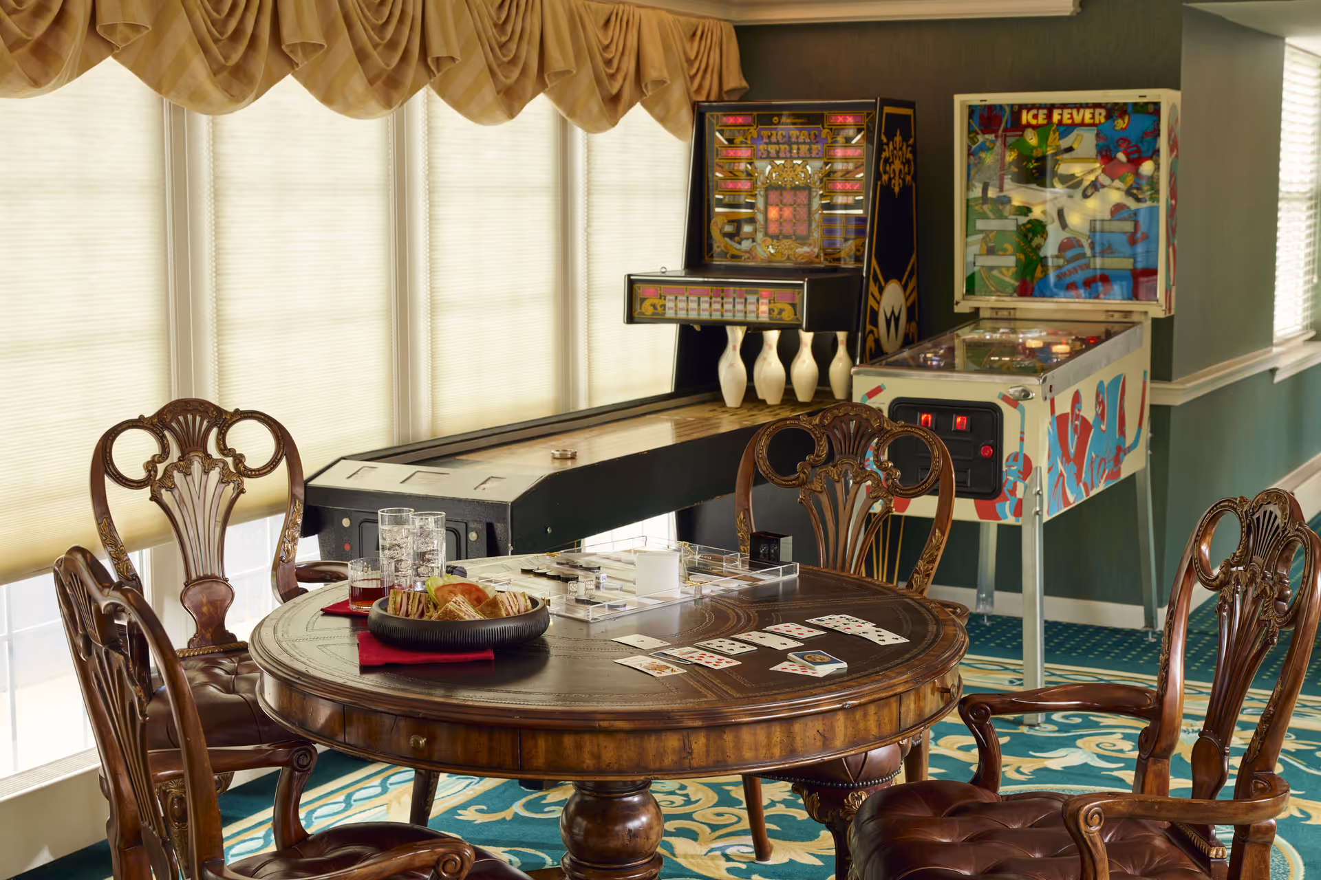 A cozy game room with a round wooden table surrounded by four ornate wooden chairs. On the table are playing cards, a board game, a plate of sandwiches, and several glasses. In the background, there is a vintage shuffleboard table with bowling pins and a pinball machine with colorful artwork. The room has large windows with beige curtains and a patterned carpet.