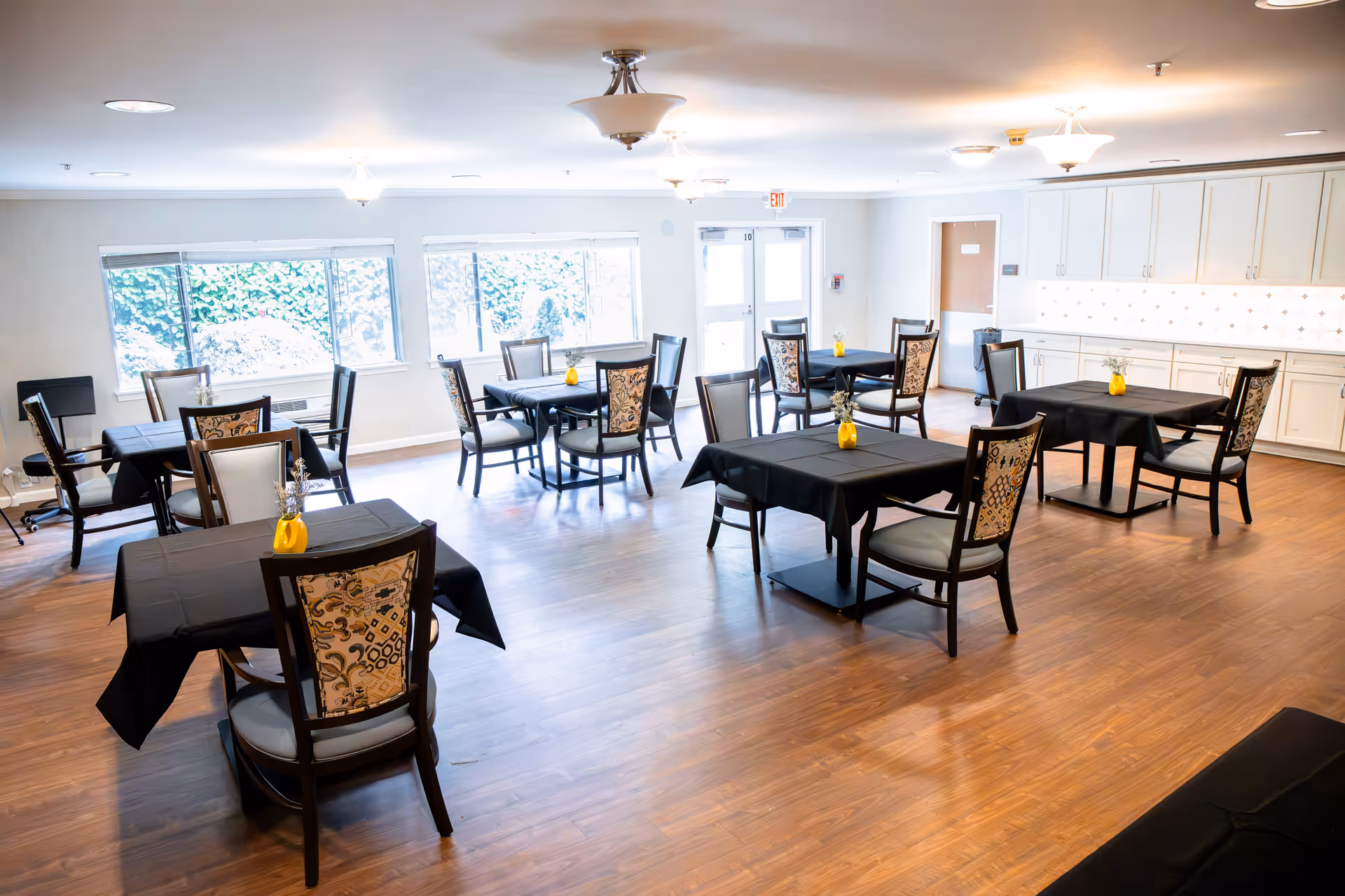 A bright dining room with several tables covered in black tablecloths, each with a small yellow vase holding flowers. The room has large windows letting in natural light, wooden flooring, and white cabinets along one wall.